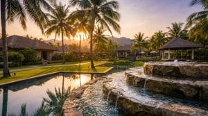 An elegant resort water feature at golden hour: a tiered stone fountain beside a still reflecting pond in a manicured tropical landscape, framed by palms and lush planting, clear sparkling water, a luxury Southeast Asian hospitality setting, warm inviting light, no people.